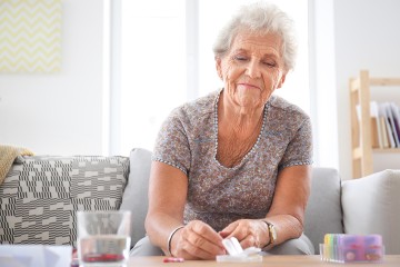 eine Seniorin sitzt an einem Tisch, vor ihr verschiedene Tablettenschachteln, die sie anfasst