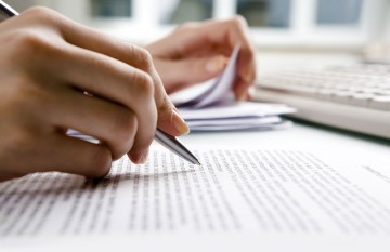 Eine Hand mit Stift und bedrucktem Papier sowie eine Tastatur.