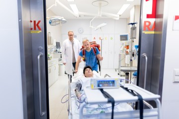 ein Sanitäter schiebt ein Krankenbett mit einem jungen Patienten aus einem Raum im Krankenhaus, im Hintergrund schaut ein Arzt hinterher