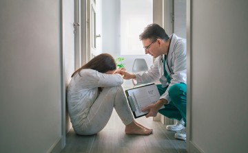 Ein Arzt hockt vor einer Patientin, die niedergeschlagen am Boden sitzt. Er berührt ihren Arm. 