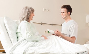 Das Bild zeigt eine Patientin im Krankenhaus und einen Pfleger, der ihr vorliest.