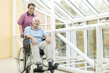 Älterer Mann im Rollstuhl wird von einer Pflegerin geschoben.
