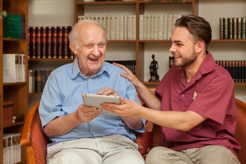 Ein junger Pflege sitzt neben einem älteren Mann und erklärt etwas anhand eines Tablets.