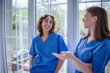 zwei junge Pflegerinnen im blauen Kittel stehen an einem Fenster und unterhalten sich