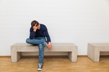 Ein junger Mann sitzt auf einer Bank und stützt den Kopf mit einer Hand.