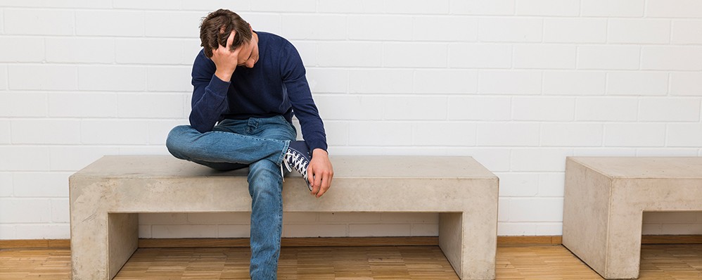 Ein junger Mann sitzt auf einer Bank und stützt den Kopf mit einer Hand.