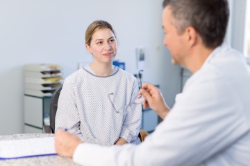 Eine junge Frau in einem OP-Oberteil sitzt vor einem Arzt, der ihr etwas erklärt.